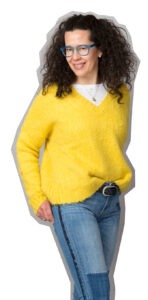 Stehende junge Dame in einem gelben Pullover mit V-Ausschnitt, Jeans mit Pailletten an den Seiten, offenem Haar und Brille mit blauer Fassung